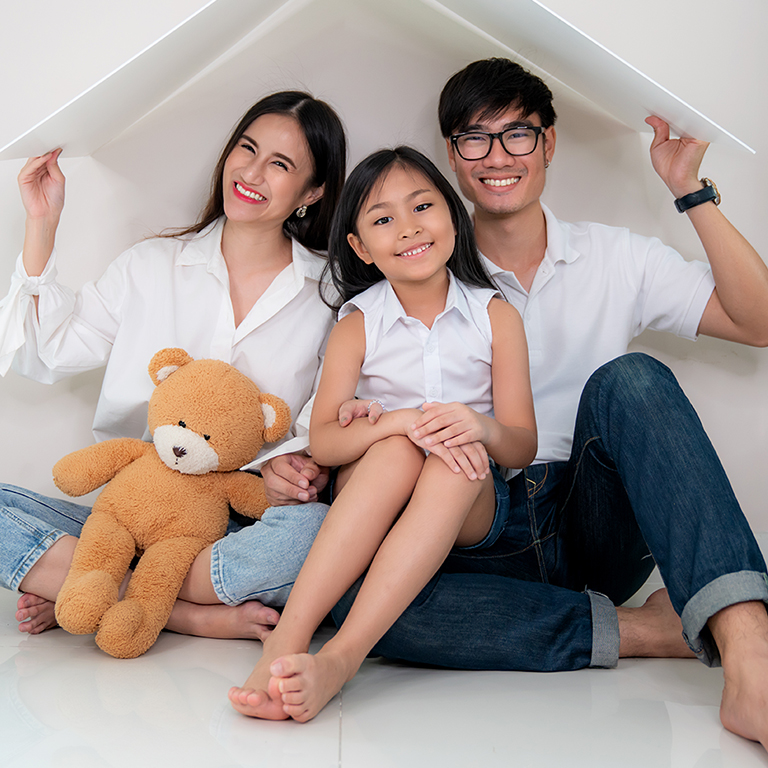 Asian Young parents keeping their hands close forming roof over their daughter while sitting on the floor of living room at home. Concept of housing for young family.